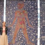 A student stands in front of the artwork she created, that features a red coloured figure on a blue patterned background.