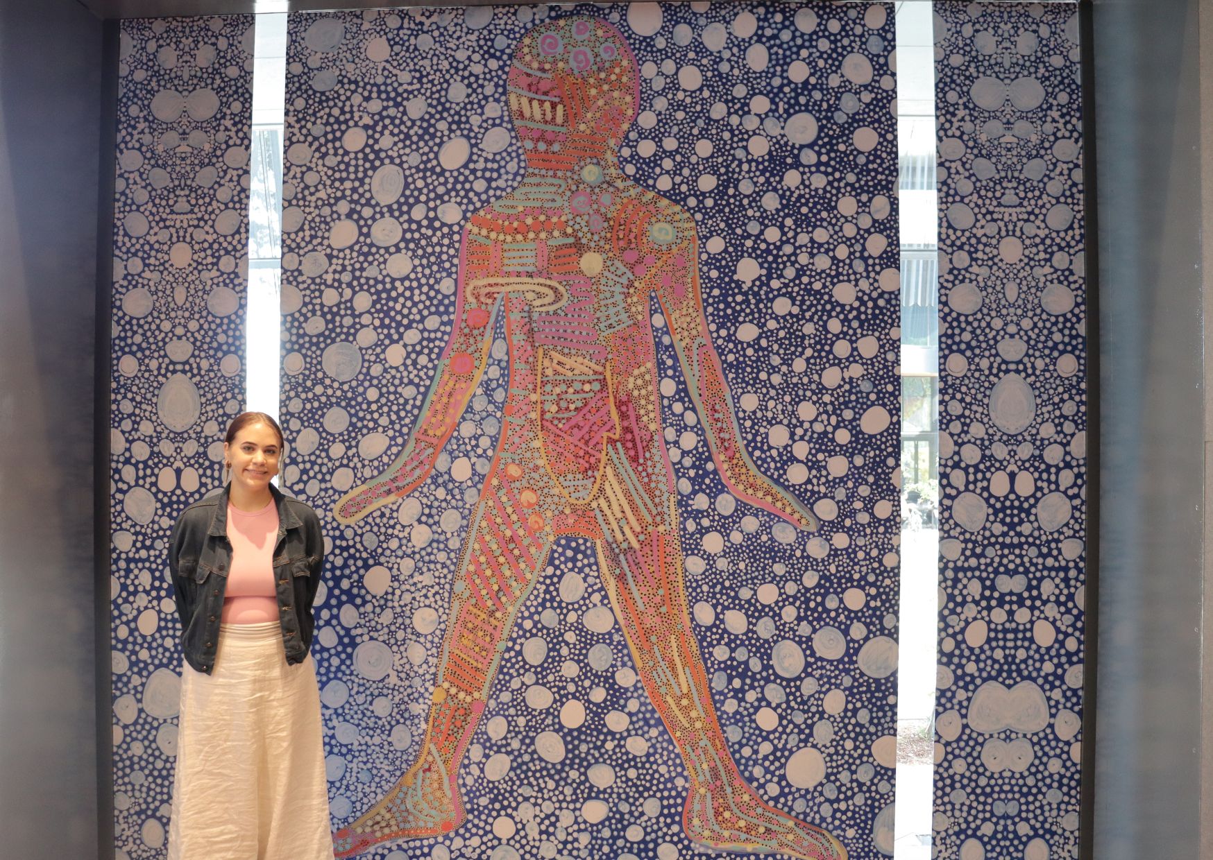 A student stands in front of the artwork she created, that features a red coloured figure on a blue patterned background.