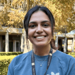 A student stands smiling on campus, in front of a building and trees.