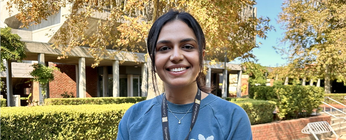 A student stands smiling on campus, in front of a building and trees.