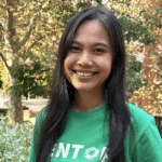 A student stands smiling with the hair down. She wears a green shirt that says 'Mentor' on it. Behind her there are trees and a building by her right shoulder.