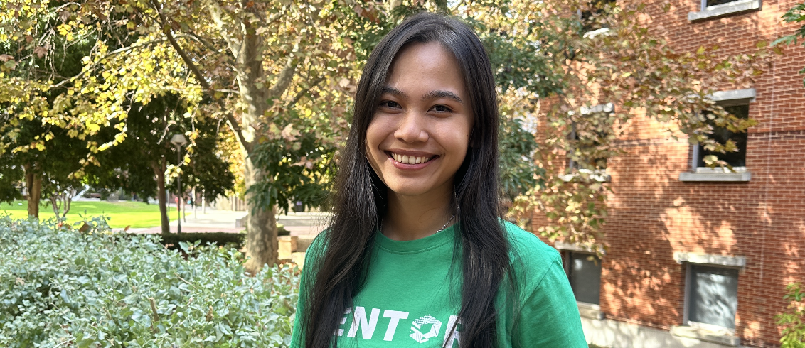 A student stands smiling with the hair down. She wears a green shirt that says 'Mentor' on it. Behind her there are trees and a building by her right shoulder.