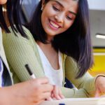 Two women looking down at notes, one smiling, one writing with a pen.