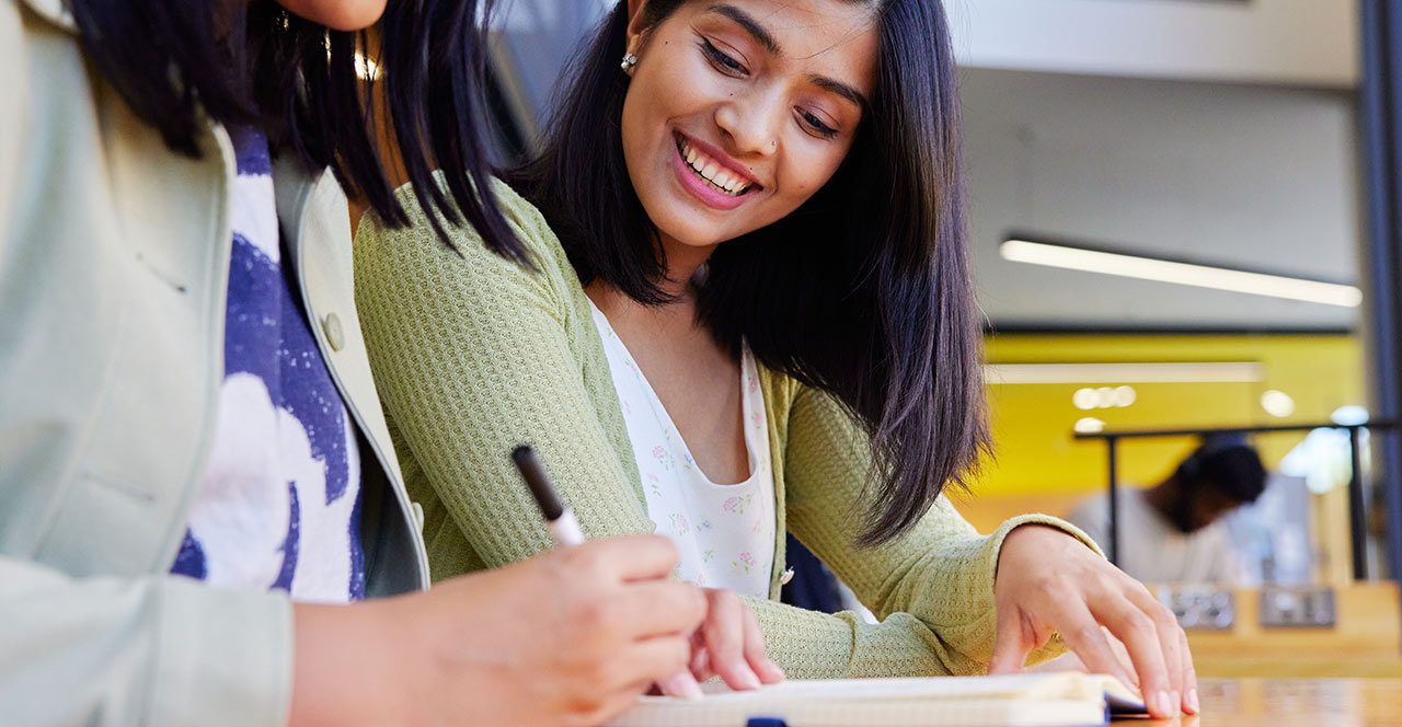 Two women looking down at notes, one smiling, one writing with a pen.