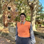 Brooke stands smiling on campus, wearing a bright orange vest with a long sleeve, collared shirt.