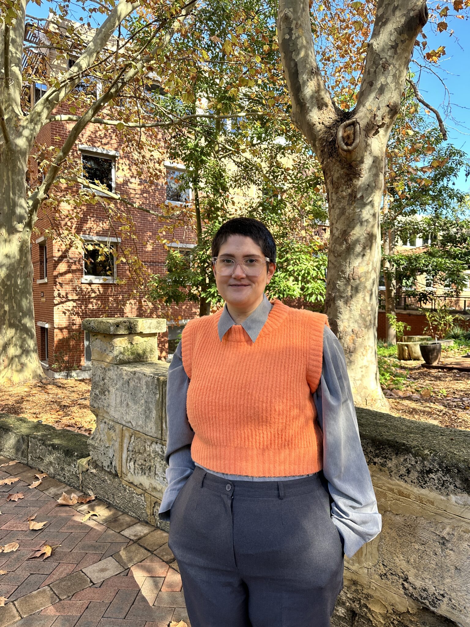 Brooke stands smiling on campus, wearing a bright orange vest with a long sleeve, collared shirt.