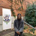 A student, Ortinel, stands smiling in front of some art and greenery.