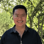 Nathan stands smiling in front of greenery. He wears his glasses and a dark collared shirt.