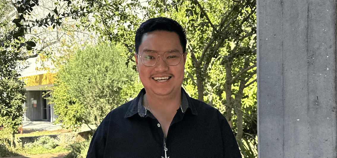 Nathan stands smiling in front of greenery. He wears his glasses and a dark collared shirt.
