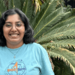 Namishka stands smiling in a blue shirt. She stands in front of a green plant.