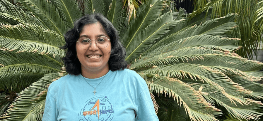 Namishka stands smiling in a blue shirt. She stands in front of a green plant.
