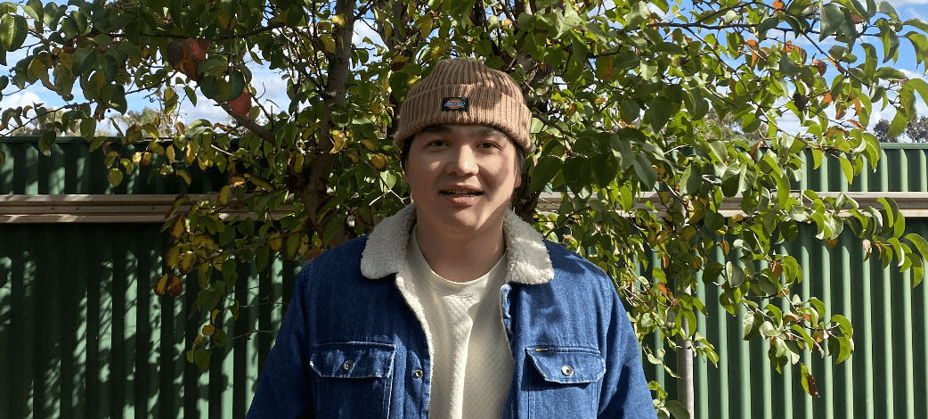 Ronnie stands smiling in front if a green fence and tree.