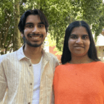 Aadhikar (Aadhi) And Akshita stand next to each other smiling, in front of a tree and building.