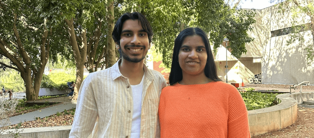 Aadhikar (Aadhi) And Akshita stand next to each other smiling, in front of a tree and building.