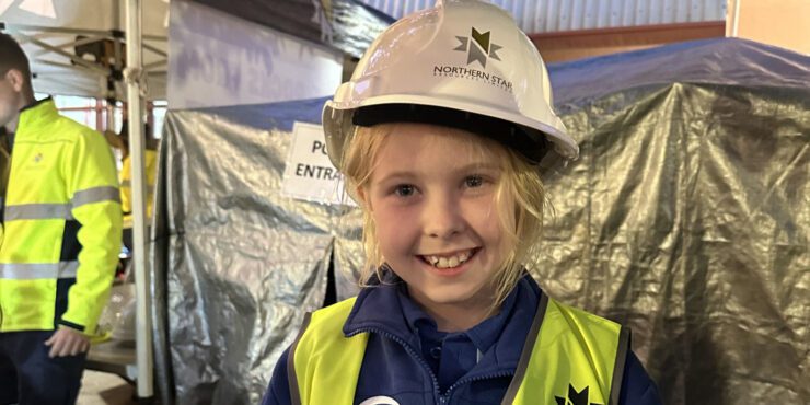 A child smiles at the camera wearing a hard hat and hi-vis vest.
