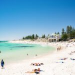 A white sandy beach with aquamarine waters. A white historical building overlooks the ocean.