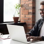 Young professional male, smiling, sitting at a desk at home and working on a laptop.
