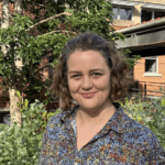 Laura stands smiling with a colourful, patterned shirt on, in front of greenery.