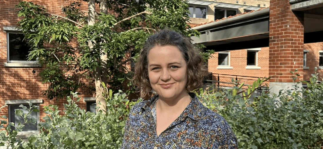 Laura stands smiling with a colourful, patterned shirt on, in front of greenery.