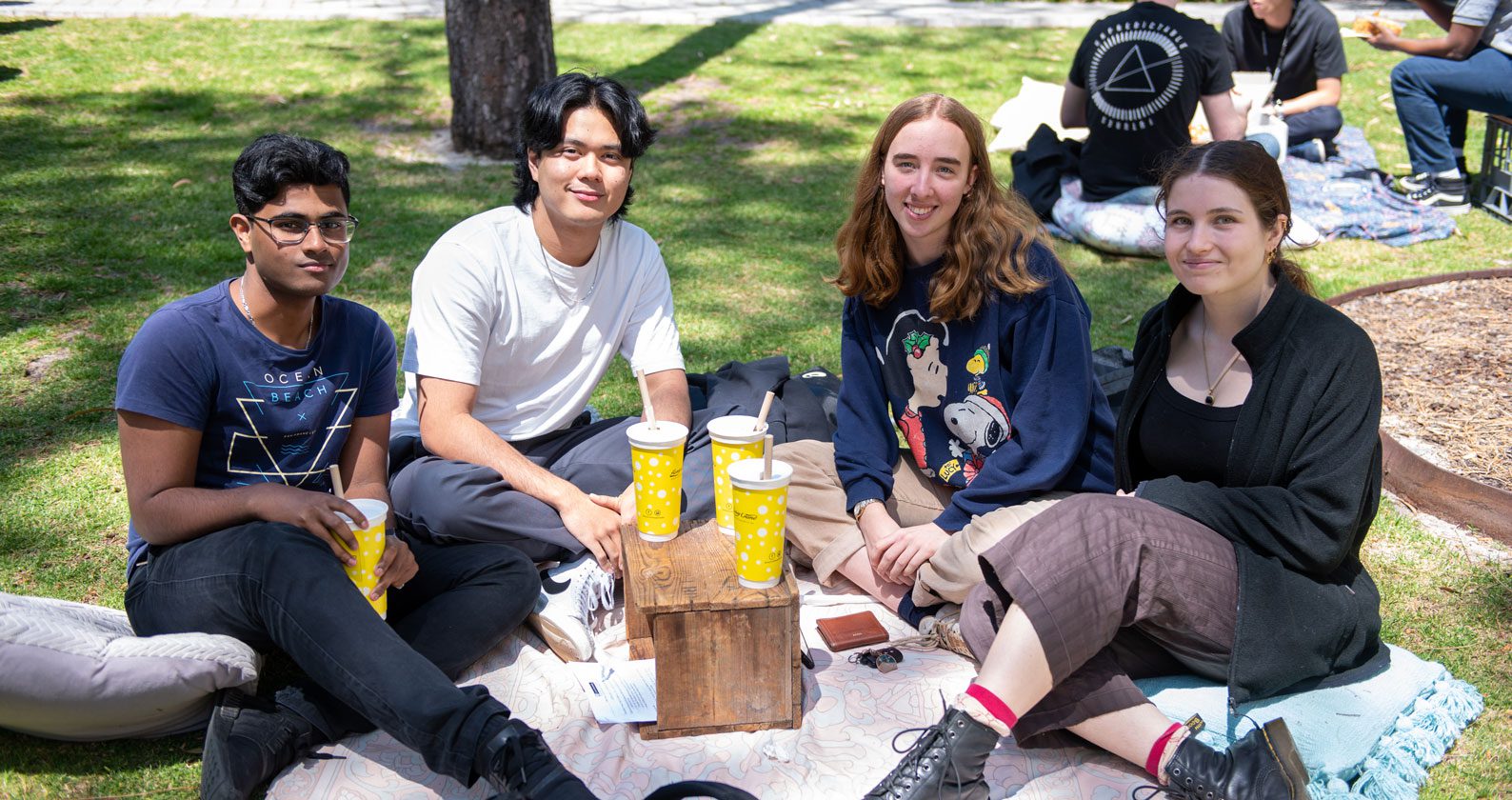Four student sit on a rug at the Creative Quarter around a table with bubble tea. They smile at the camera.