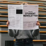 Person standing in a lecture theatre holding a newspaper in front of their face.