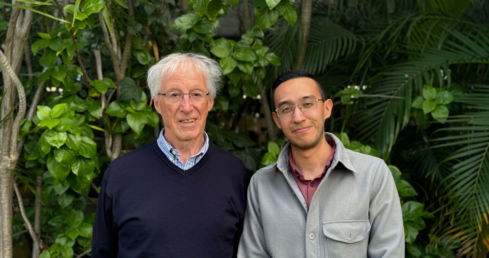 David and Bruno smile at the camera. They stand in front of a wall of plants.