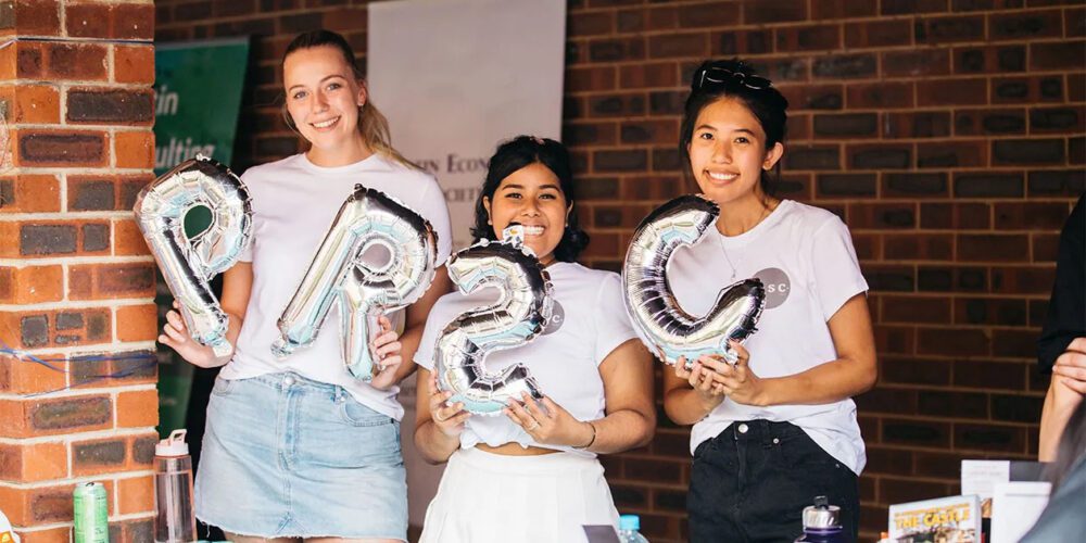 Three Curtin university students smiling and holding balloons as they enjoy an activity on campus. 