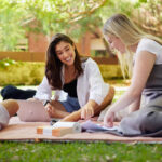 Two young women sit on a picnic blanket smiling and looking at a page of notes with an open laptop in front of them; a young male is also sits on the blanket on the edge of the image.