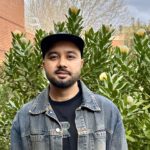 Success, a Master of Design student smiles for a photo wearing a denim jacket and black cap. Whilst standing in front of a pincushion bush.