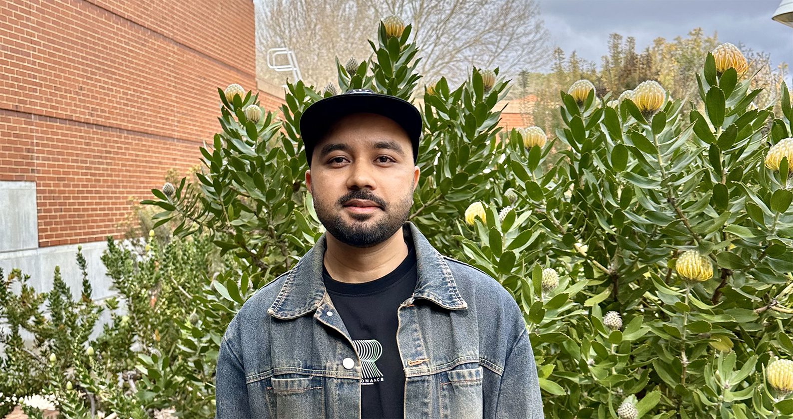 Success, a Master of Design student smiles for a photo wearing a denim jacket and black cap. Whilst standing in front of a pincushion bush.