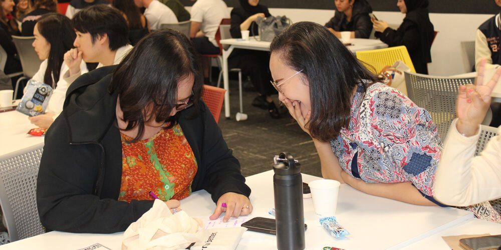 Two Faculty of Business and Law students laugh together whilst sitting down at a table to do activities.