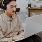 A male student wearing headphones, sitting in an armchair, and typing on a laptop.