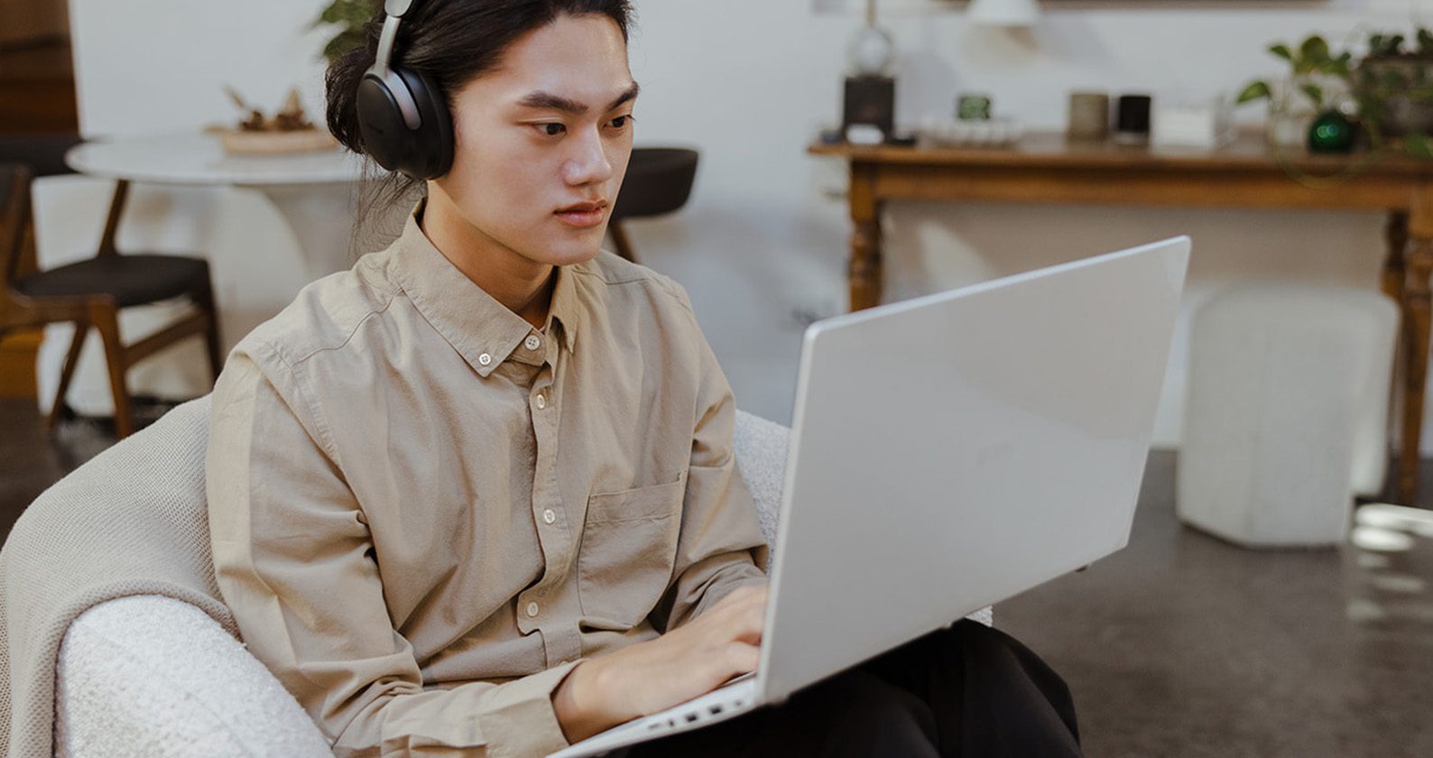 A male student wearing headphones, sitting in an armchair, and typing on a laptop.