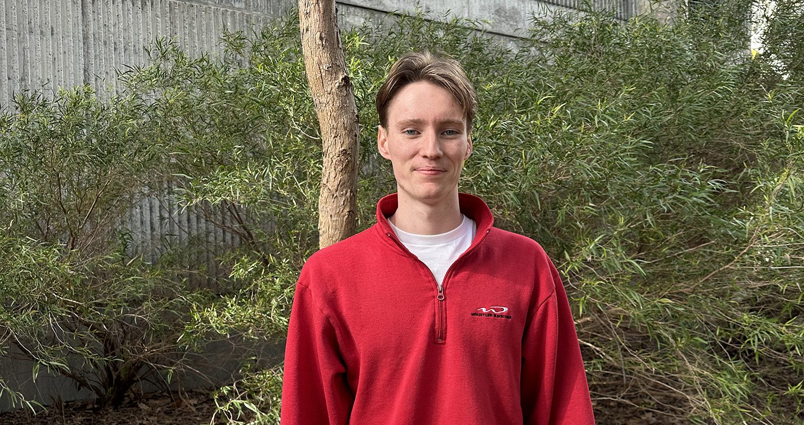 Pierce stands smiling at the camera, he wears a red jumper with a white t-shirt underneath. He is standing outside in front of foliage and trees.