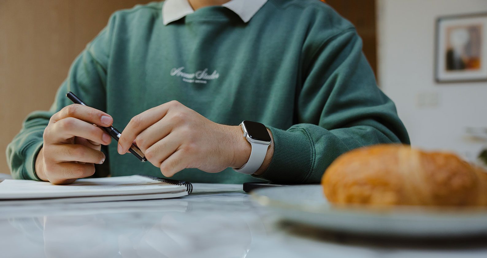 A student holding a pen above a notebook. There is a croissant to the side of the image.