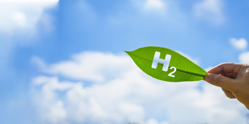 'H2' cut out from a leaf, which is held against a blue and lightly cloudy sky. This clean hydrogen energy concept demonstrates the green energy industry and alternative energy opportunities to achieve our net zero goals.