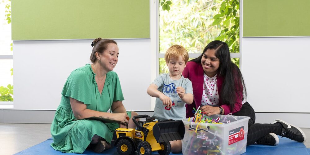 Woman and social work student sitting on a mat and smiling as a child plays with toys between them.