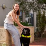 A person smiling, seated on a concrete ledge next to a black Labrador wearing a guide dog harness, in a sunny outdoor setting.