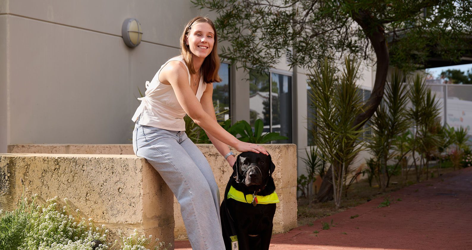 A person smiling, seated on a concrete ledge next to a black Labrador wearing a guide dog harness, in a sunny outdoor setting.