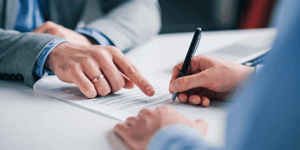 A tightly cropped image of a person is signing a piece of paper. The person opposite them is pointing out where to sign.