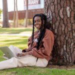 A student sitting on the grass, leaning on a tree with her laptop open.