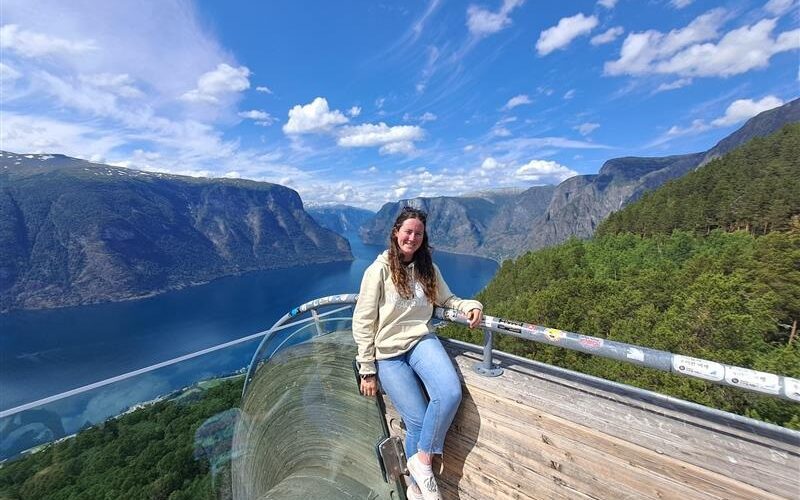 A student smiles while sitting on a wooden platform overlooking a dramatic landscape, surrounded by mountains and blue sky.