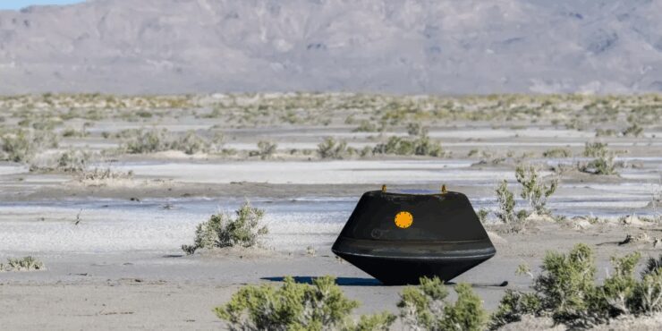 The OSIRIS-REx sample return capsule is small and black, resting on a flat, dry desert landscape with sparse bushes.