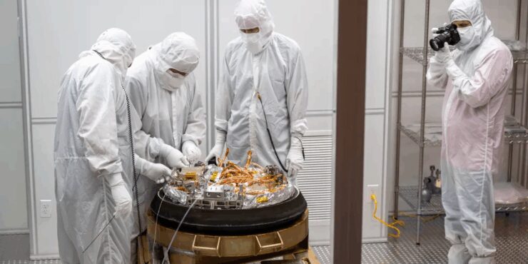 Four scientists in white cleanroom suits and gloves examine and open the the OSIRIS-REx sample return capsule. The open object is covered in wiring and sensors. A fifth person photographs the scene from the side.