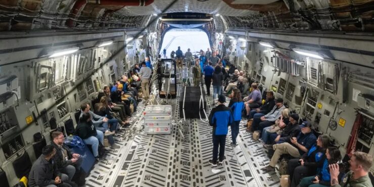 Interior of a large military cargo aircraft with people seated along both walls, some in casual clothing and others in uniforms, as crew members walk down the central aisle toward the open rear hatch.
