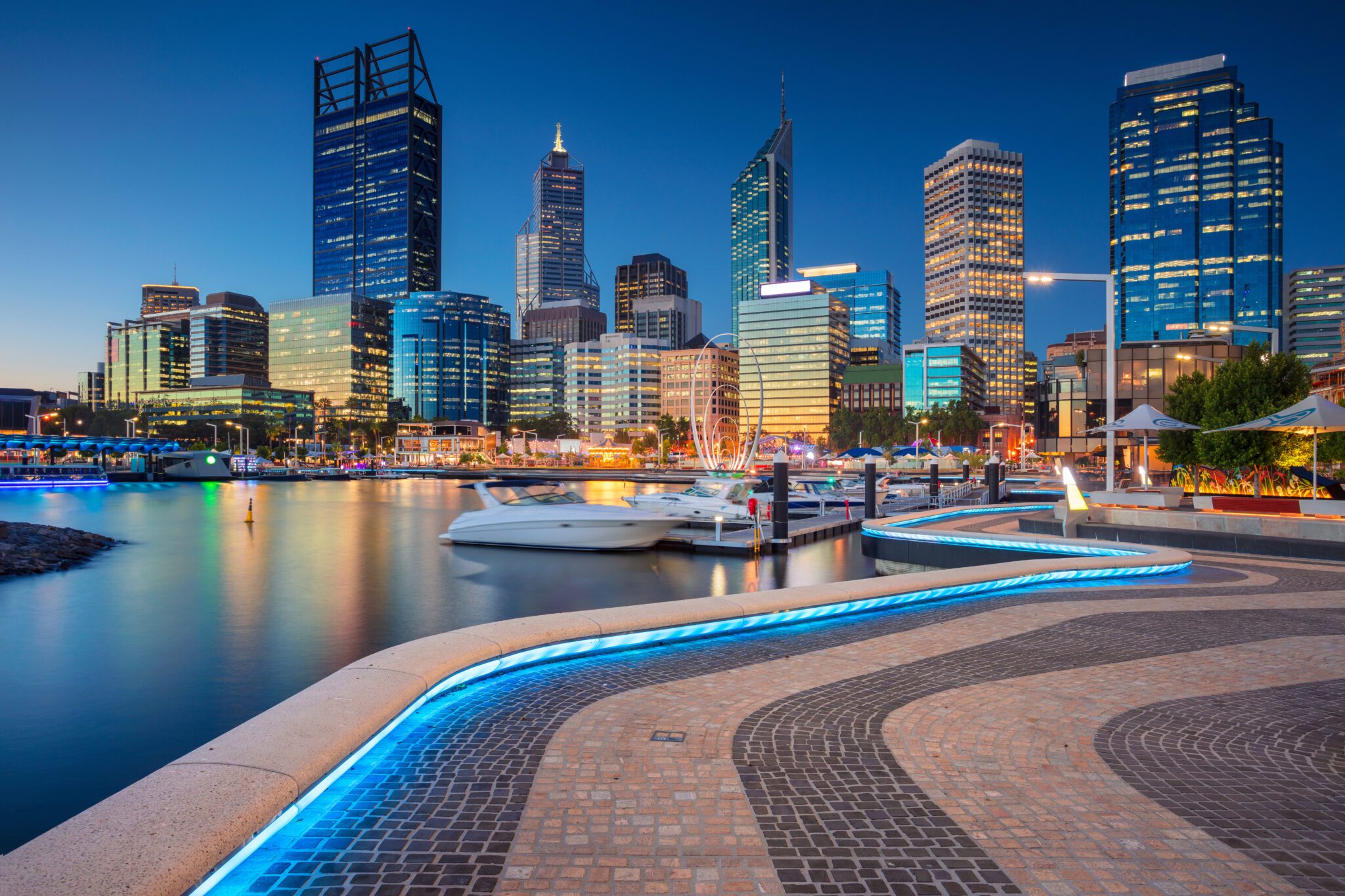 Perth city skyline at dusk, with high-rise buildings lit up and reflections on the water at Elizabeth Quay.