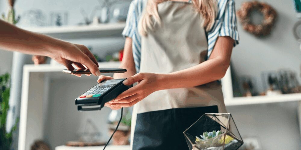 Person selling goods at a light-filled store. Cropped image of a customer paying using smart phone.