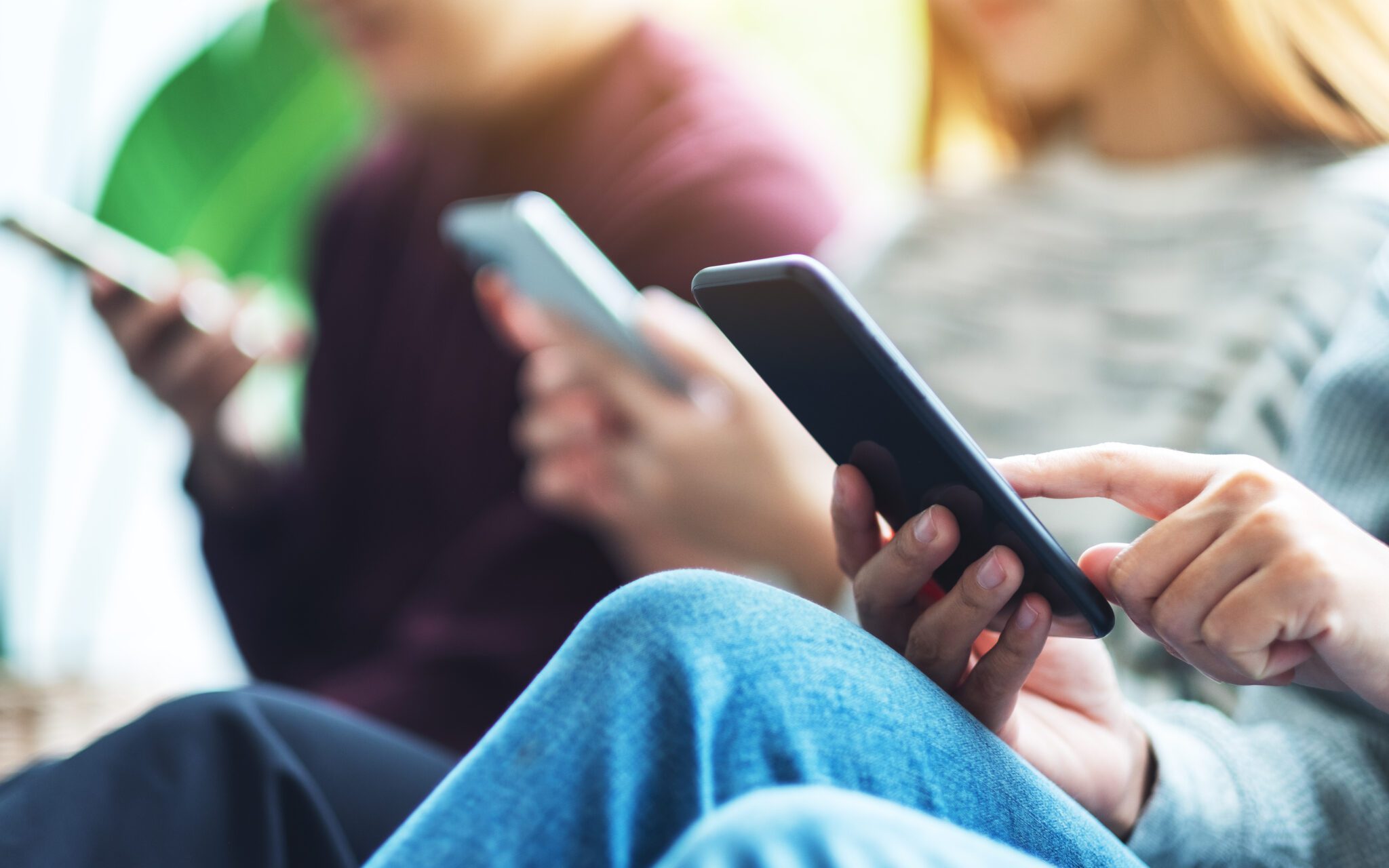 Three people sitting closely together, each holding and using a smartphone, with a blurred background