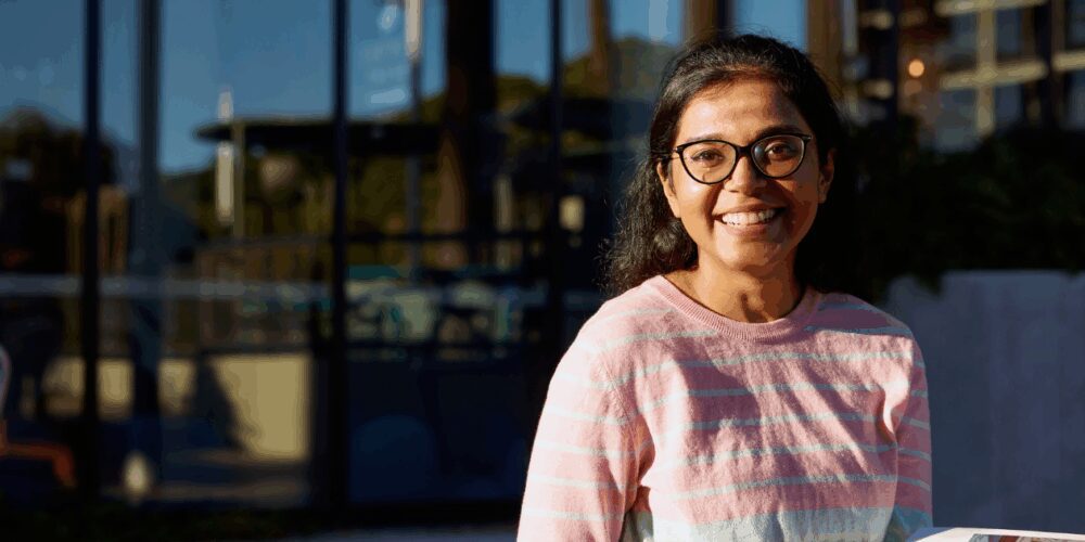 Woman in glasses and a pink striped shirt sits outside as she smiles to camera.