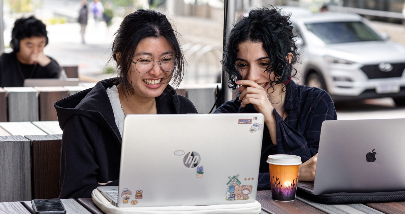Two people working on laptops at an outdoor table with a coffee cup